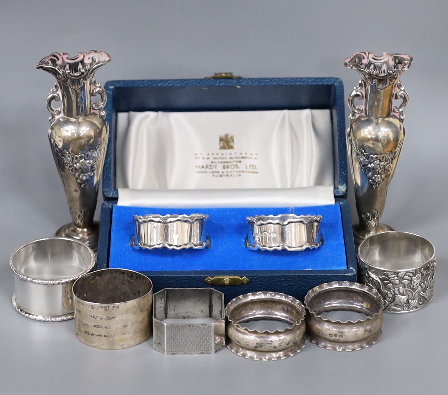 A cased pair of silver napkin rings, five others including one white metal and a two small silver vases, weighted.